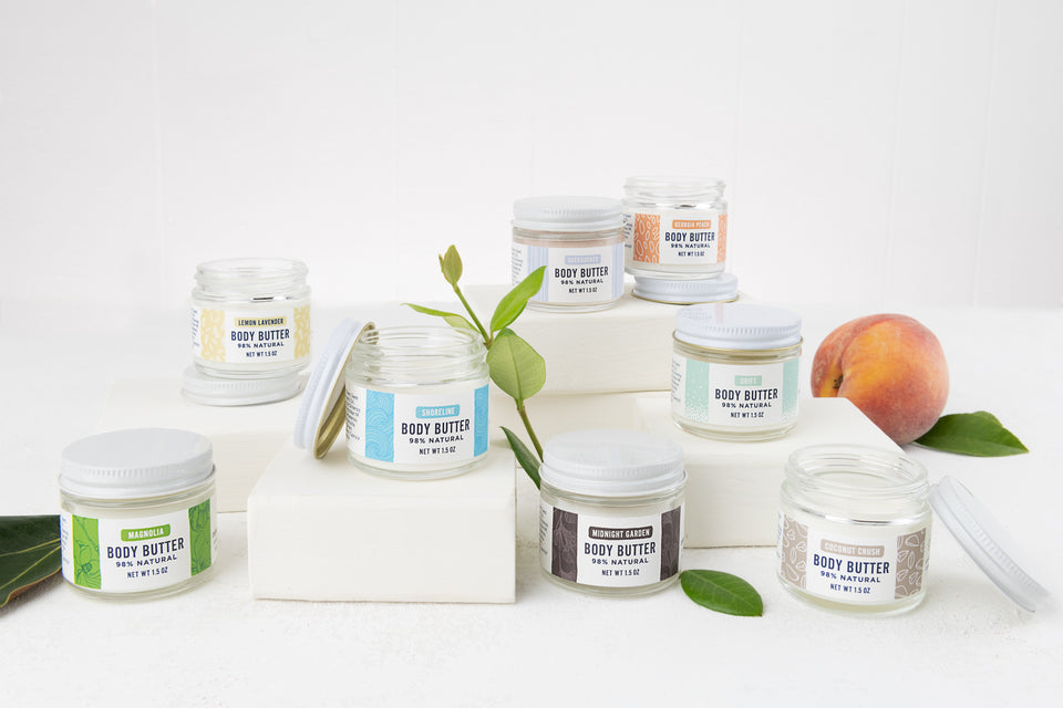 a collection of 8 small jars of body butter on a white background with sprigs of leaves and a peach